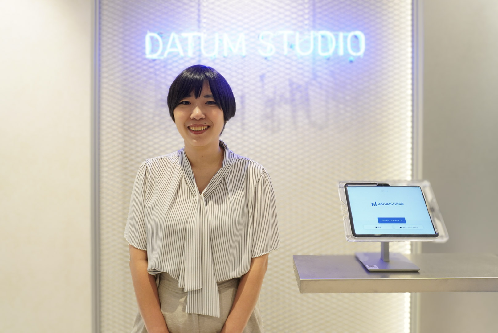 DATUM STUDIOのオフィス受付前に立つ真嘉比愛。背景にDATUM STUDIOのロゴと受付タブレットが写っている