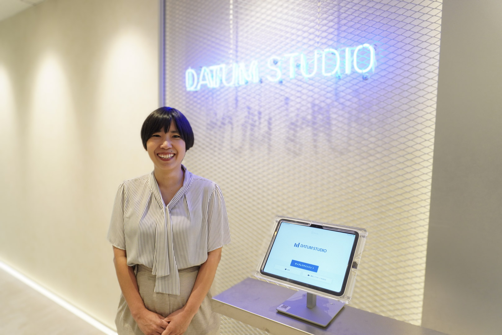 DATUM STUDIOのオフィス受付前で笑顔を見せる真嘉比愛。背景にDATUM STUDIOのロゴと受付端末がある